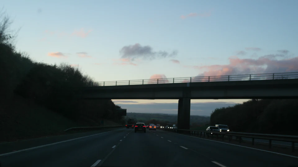 A motorway at dusk from the driver’s seat, a bridge ahead and tail lights in the distance