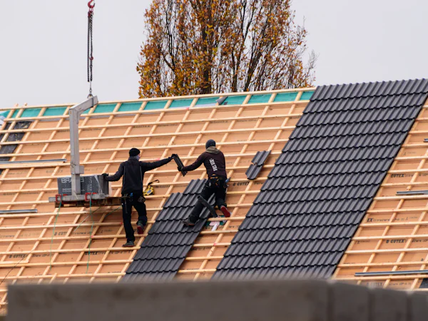 Two roofers far off on a battened roof laying dark tiles from a hoist