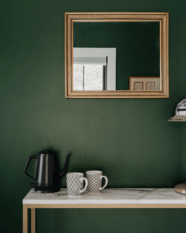 A deep green wall with a gilt mirror over a marble-topped side table