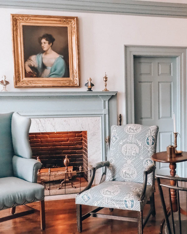A pale period sitting room with a fireplace, a portrait in a gilt frame and a grey-blue panelled door