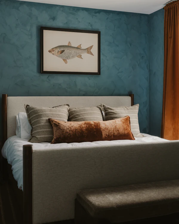 A bedroom with textured blue-green walls, a framed fish print and rust curtains