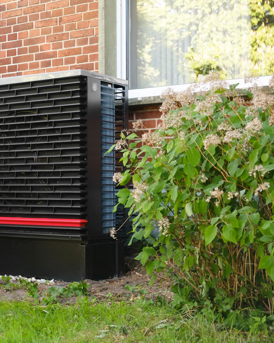 An air source heat pump beside a brick house and a flowering shrub