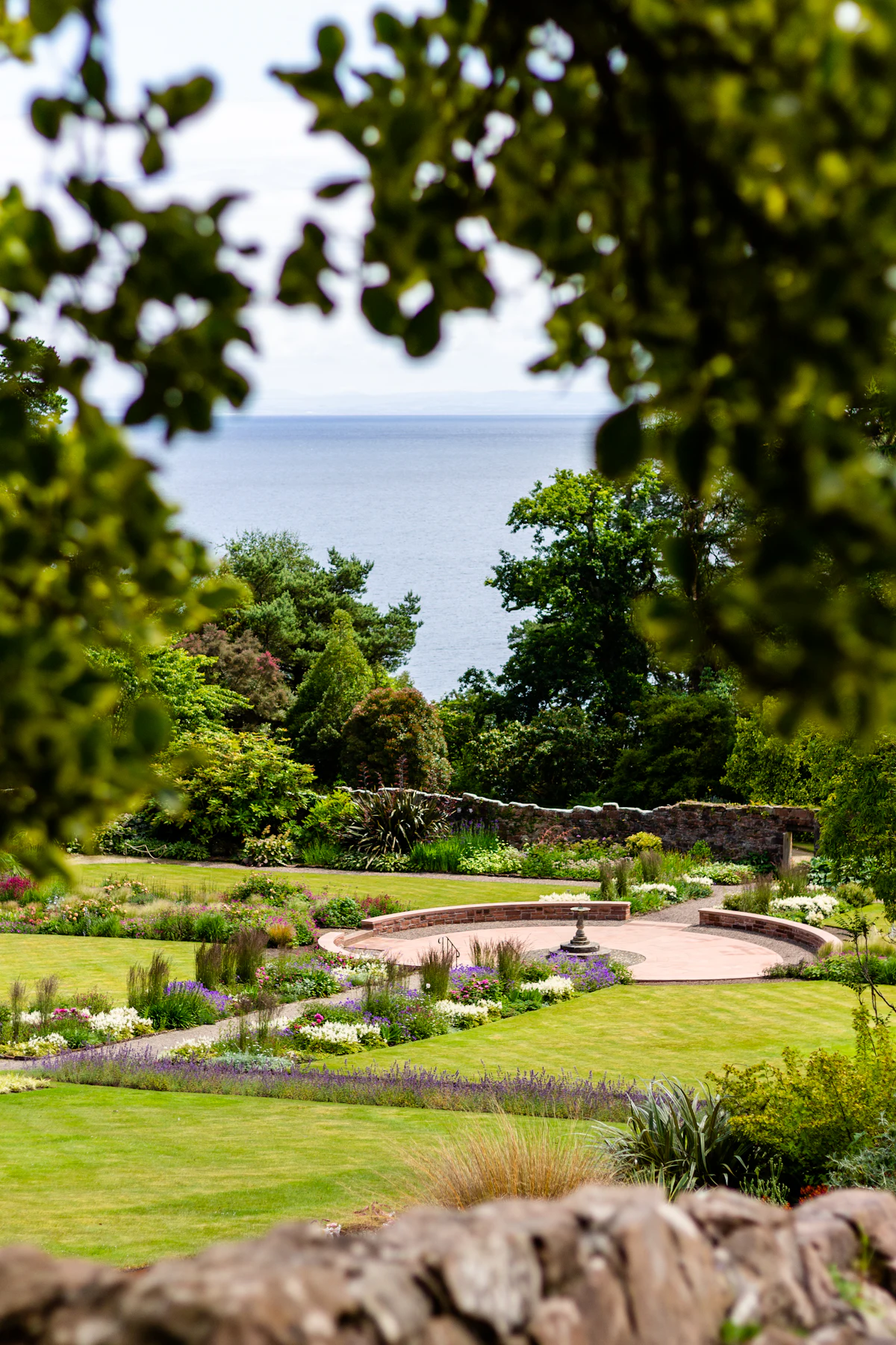 A terraced garden seen through trees: a round sunken terrace and pool below a stone wall, lawns and borders, and the sea beyond