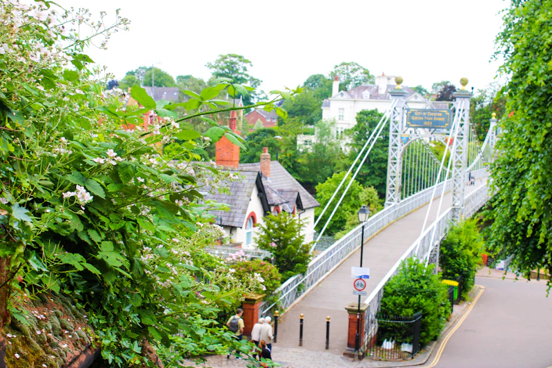 A white suspension footbridge leading over the river to villas among trees on the far bank