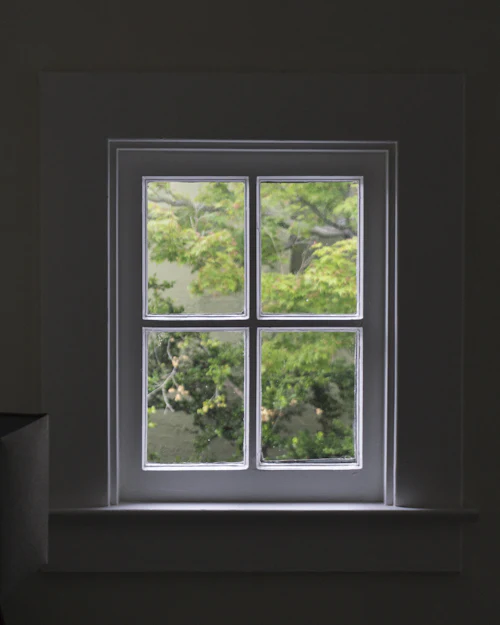 A small grey-painted cottage window from inside, green leaves outside the four panes