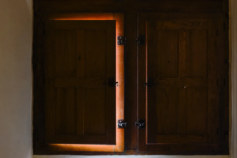 A pair of dark panelled internal shutters, one folded a little open with warm light at its edge