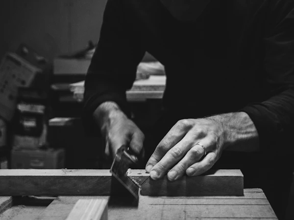 Two hands steadying a length of timber against a saw fence, in black and white