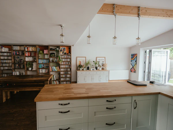 A cream kitchen peninsula with an oak worktop, a dining table and bookshelves beyond