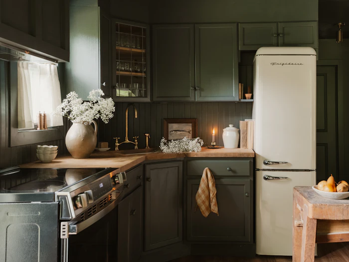 A dark green painted kitchen with oak worktops, a cream fridge and white flowers by the window