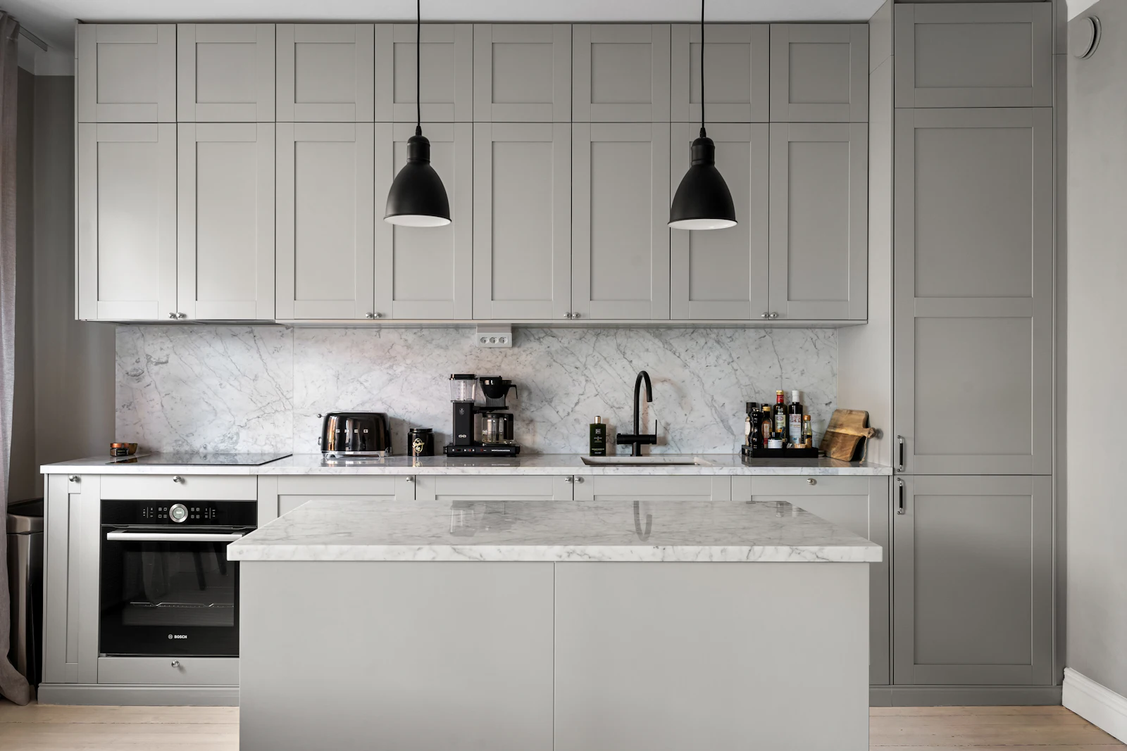 A painted grey shaker kitchen seen square-on: eight tall wall units, a larder, an oven housing and a marble-topped island under two black pendants