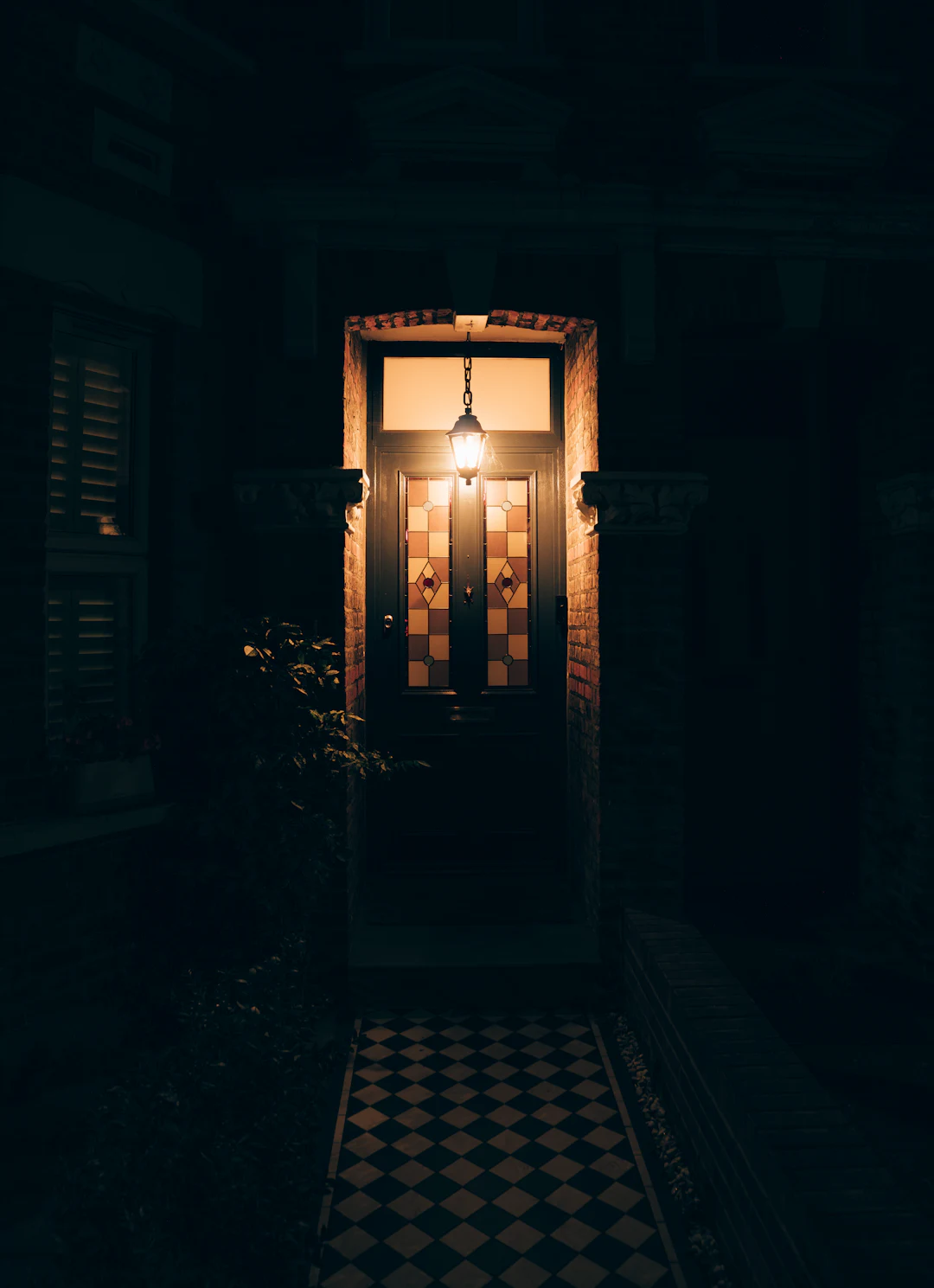 A terraced house front door at night, stained glass lit by a hanging lantern, a black and white tiled path leading up to it