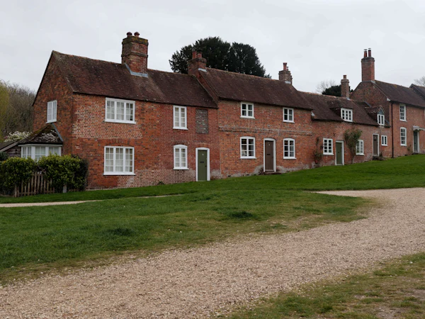 A row of old red brick cottages with small white windows behind a green