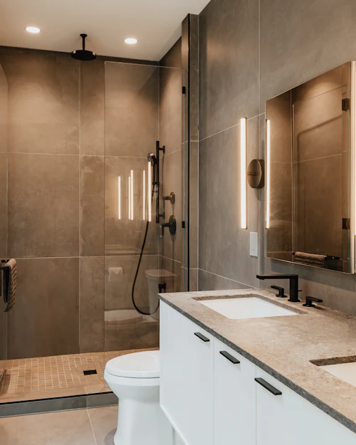 A walk-in shower in warm brown porcelain beside a vanity with lit mirrors