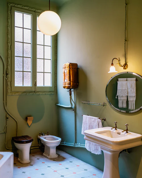 A period bathroom with sage green walls, a tall sash window, a pedestal basin and a round mirror