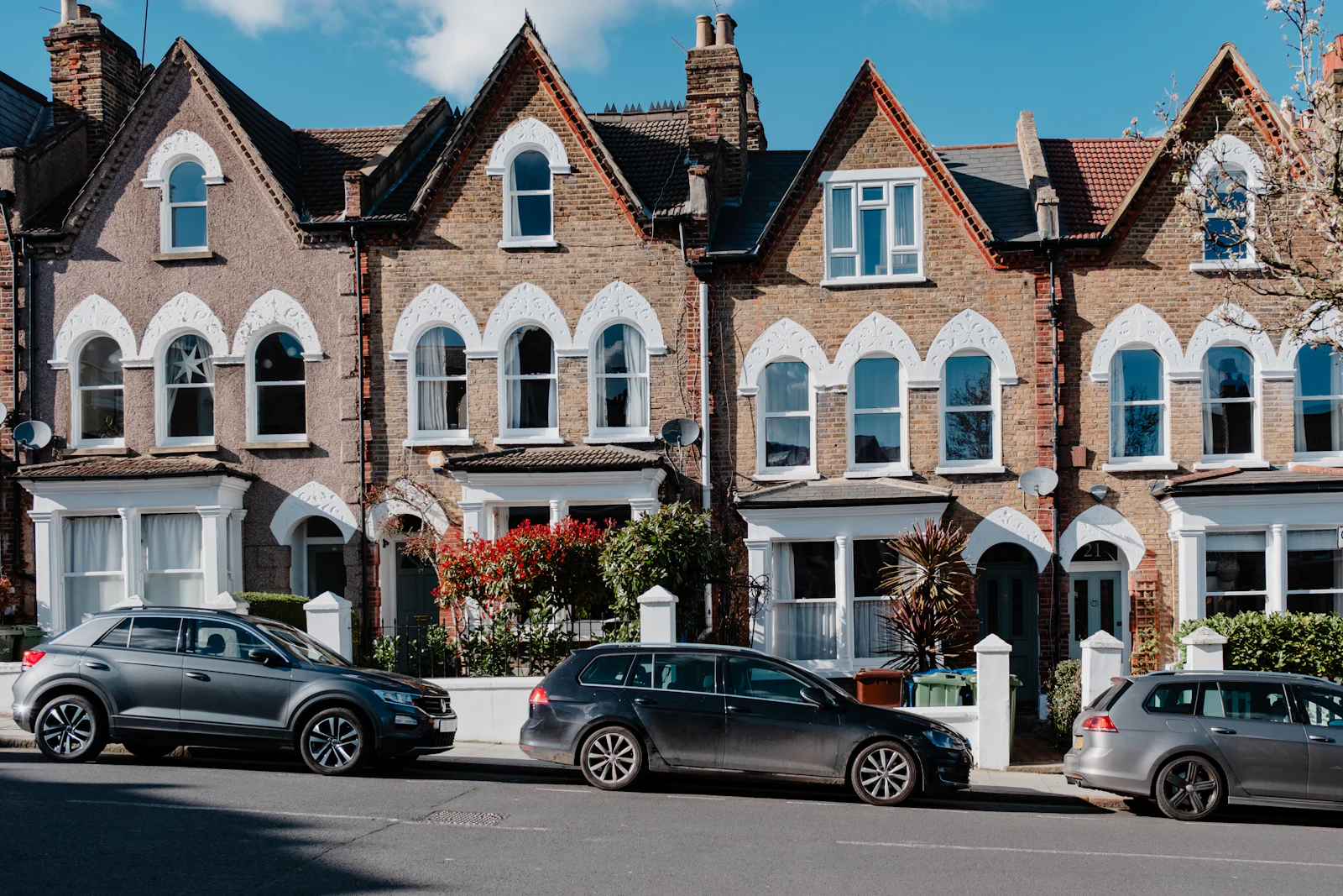 A row of gabled Victorian brick houses with bay windows and cars parked outside, under a blue sky